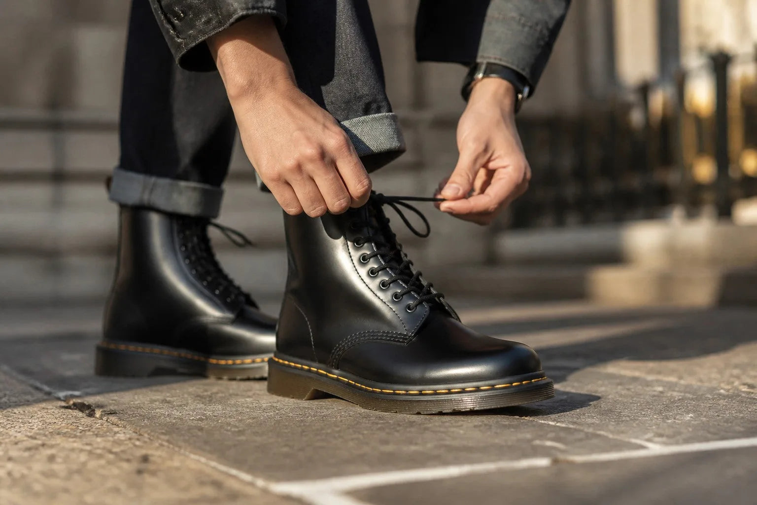 Person tying black laces on leather combat boot while standing on stone pavement