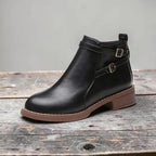 Side view of black faux leather ankle boot with double buckle straps and contrast brown sole on a wooden surface