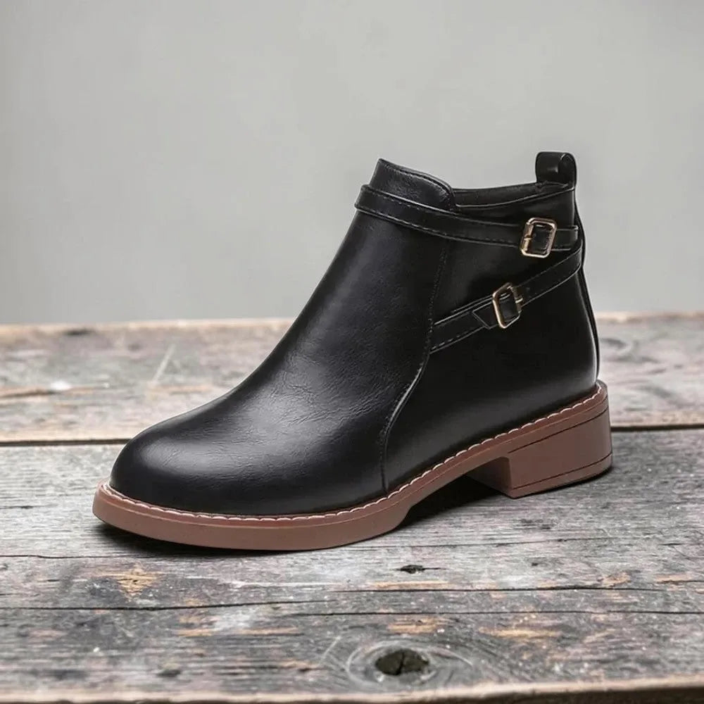 Side view of black faux leather ankle boot with double buckle straps and contrast brown sole on a wooden surface