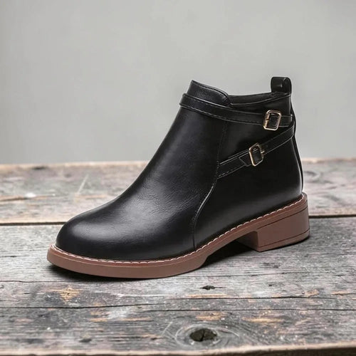 Side view of black faux leather ankle boot with double buckle straps and contrast brown sole on a wooden surface