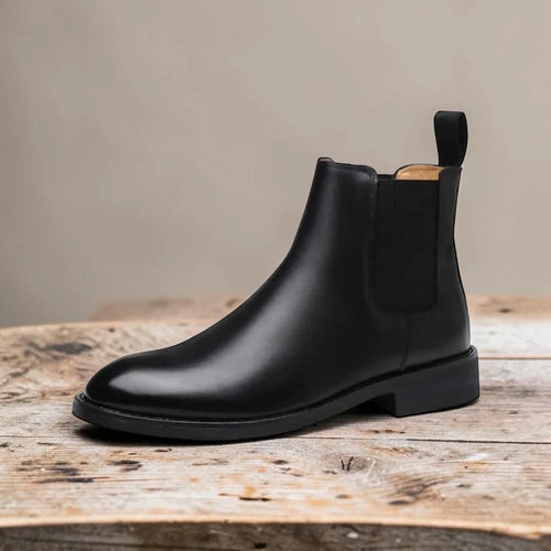 Side profile of a black leather Chelsea boot with elastic gusset and pull tab, resting on a wooden table