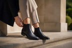 Close-up of a hand adjusting the pull tab on a black leather Chelsea boot outdoors in sunlight