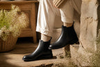 Woman seated wearing black leather Chelsea boots with cream trousers and knit socks near a wicker basket of flowers